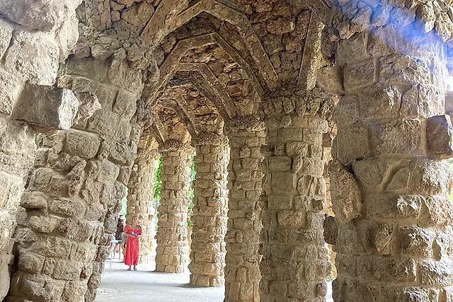 A viaduct of Park Güell: rough stone columns leaning like tree trunks under a vaulted stone walkway, with greenery behind